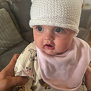 Leila joined the competition — help win amazing prizes! baby, infant, hat, knitted_hat, pink_bib, cute, wide_eyes, expression, indoor, living_room, sofa, person, child, hand, soft_lighting, face, clothing, cute_expression, holding, closeup