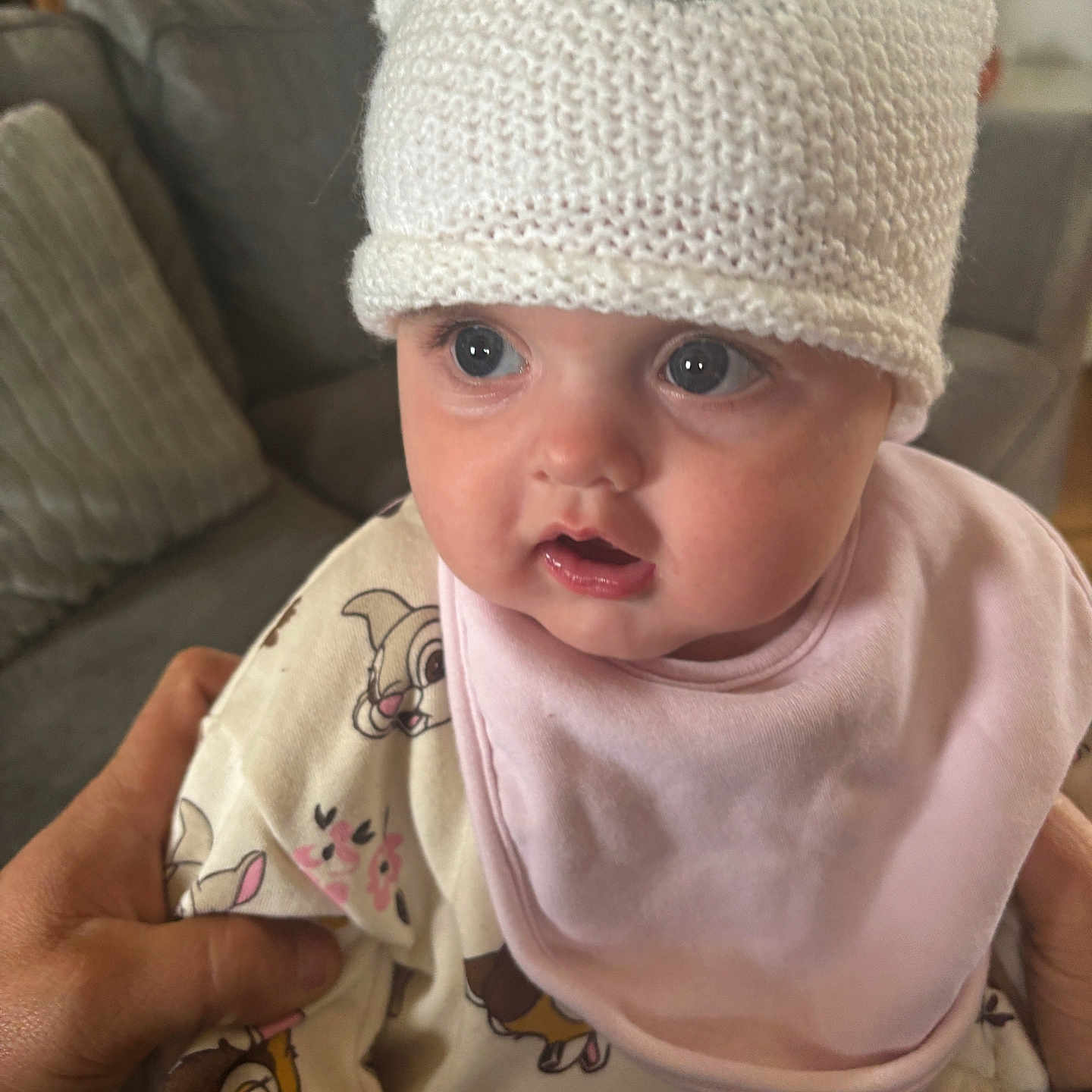 Leila joined the competition — help win amazing prizes! baby, child, closeup, clothing, cute, cute_expression, expression, face, hand, hat, holding, indoor, infant, knitted_hat, living_room, person, pink_bib, sofa, soft_lighting, wide_eyes