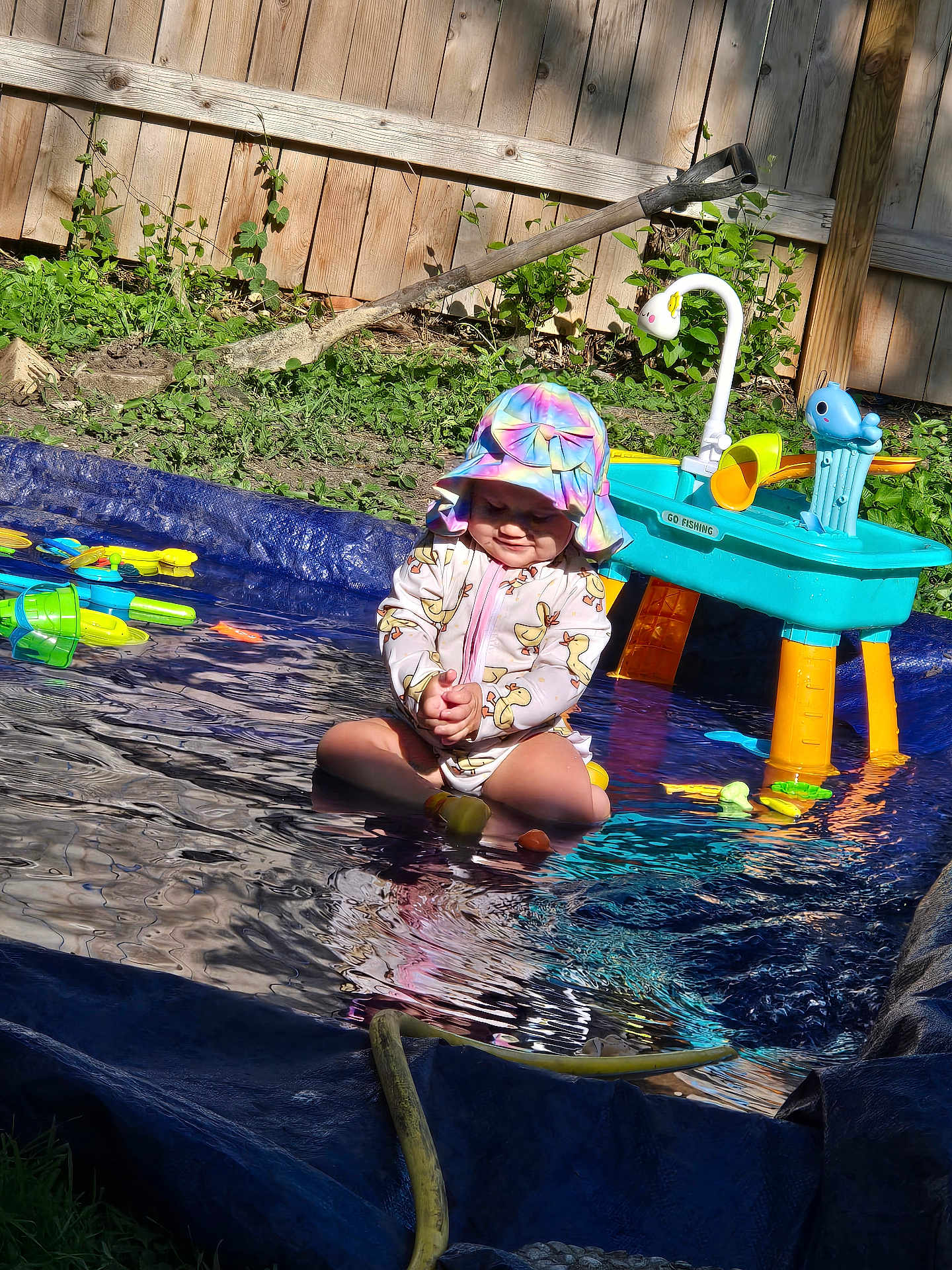 Amara is registered to the contest to win money with this photo: toddler, child, pool, water, outdoor, sunhat, swimsuit, toy, plastic, play, garden, fence, summer, sitting, water_toy, hose, sunlight, nature, grass, happy