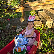Amara joined the competition — help win amazing prizes! toddler, child, pink_dress, bunny_ears, wagon, red_wagon, outdoor, greenery, sunlight, toy_set, basket, grass, stone_steps, wooden_fence, happy, smiling, person, nature, playtime, garden