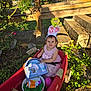 toddler, child, pink_dress, bunny_ears, wagon, red_wagon, outdoor, greenery, sunlight, toy_set, basket, grass, stone_steps, wooden_fence, happy, smiling, person, nature, playtime, garden
