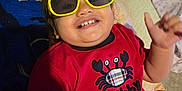 Amara is registered to the contest to win money with this photo: toddler, child, smiling, yellow_sunglasses, red_shirt, crab_design, bed, blanket, indoor, portrait, happy, cute, person, clothing, casual, fun, baby, head, face, cheerful