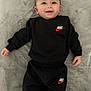 baby, smiling, black_clothing, nike_logo, infant, lying_down, blanket, cute, happy, child, face, sleeves, pants, soft_texture, portrait, young_child, indoor, casual_wear, person, comfortable