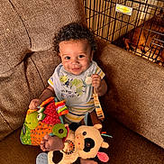 Giovanni joined the competition — help win amazing prizes! baby, child, smiling, curly_hair, bib, toy, stuffed_animal, couch, brown_fabric, dog_crate, dog, indoor, happy, playful, infant, home, pet, retriever, colorful, comfortable