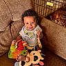 baby, child, smiling, curly_hair, bib, toy, stuffed_animal, couch, brown_fabric, dog_crate, dog, indoor, happy, playful, infant, home, pet, retriever, colorful, comfortable