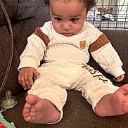 Giovanni joined the competition — help win amazing prizes! baby, bare_feet, brown_couch, child, clothing, cozy, curious, curly_hair, cute, face, home, indoor, infant, looking_down, pacifier, person, relaxed, seated, toy, white_outfit