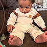 baby, bare_feet, brown_couch, child, clothing, cozy, curious, curly_hair, cute, face, home, indoor, infant, looking_down, pacifier, person, relaxed, seated, toy, white_outfit
