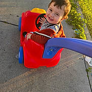 Omar joined the competition — help win amazing prizes! toddler, child, toy_car, outdoor, sidewalk, smiling, happy, sunlight, play, red, blue, plastic, fence, greenery, casual_clothing, face, person, young_child, daytime, cute