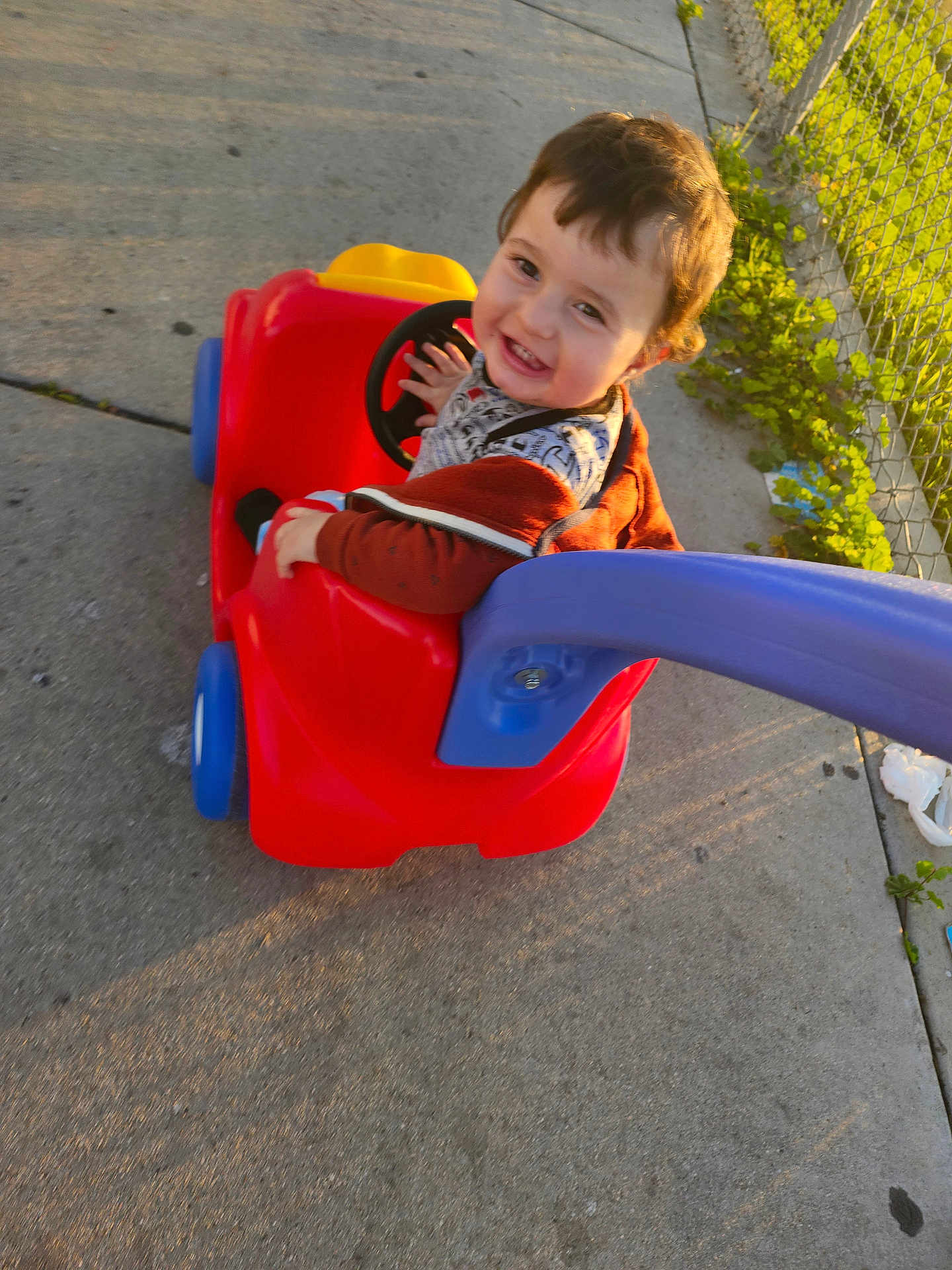Omar joined the competition — help win amazing prizes! toddler, child, toy_car, outdoor, sidewalk, smiling, happy, sunlight, play, red, blue, plastic, fence, greenery, casual_clothing, face, person, young_child, daytime, cute