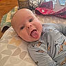 Theodore is registered to the contest to win money with this photo: baby, tongue_out, blue_onesie, airplane_pattern, cushion, table, candle, red_cloth, glass, wooden_table, indoor, playful, child, face, expression, person, home, cute, infant, smiling