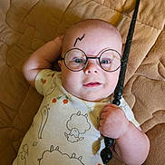 Theodore is registered to the contest to win money with this photo: baby, infant, glasses, wand, lightning_bolt, onesie, sheep_pattern, quilted_blanket, lying_down, hand, face, portrait, costume, cute, child, playful, indoor, expression, young, magic