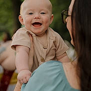Judson is registered to the contest to win money with this photo: baby, child, smiling, woman, holding, outdoor, greenery, happy, portrait, person, face, short_hair, clothing, buttoned_shirt, hand, shoulder, daylight, casual, skin, expression