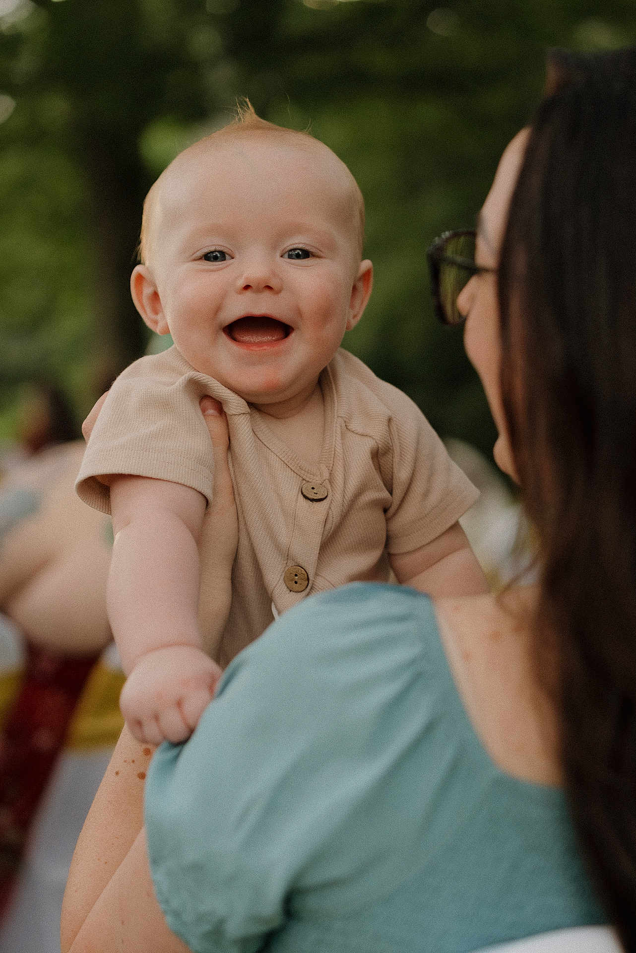 Judson is registered to the contest to win money with this photo: baby, child, smiling, woman, holding, outdoor, greenery, happy, portrait, person, face, short_hair, clothing, buttoned_shirt, hand, shoulder, daylight, casual, skin, expression