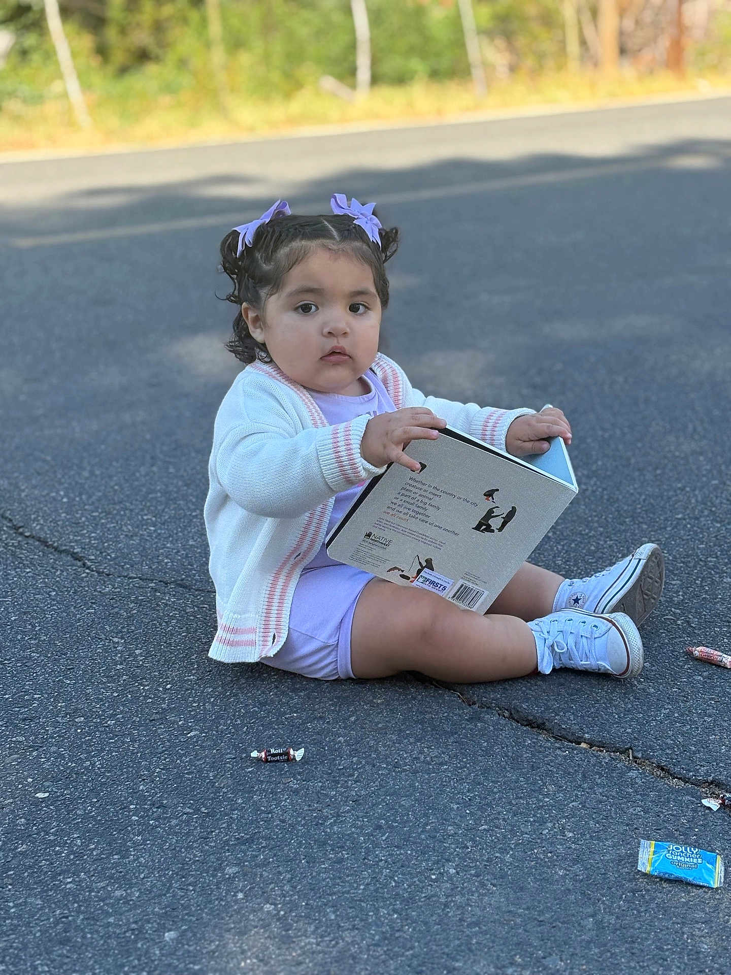 Yahzmeen is registered to the contest to win money with this photo: toddler, child, book, road, asphalt, candy, wrapper, shoes, sneakers, clothing, hair_bows, outdoor, sitting, curly_hair, white_cardigan, purple_outfit, person, daylight, cute, playful