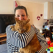 Ron is registered to the contest to win money with this photo: baby, bracelet, cat, child, decorations, furniture, holding, home, indoor, living_room, orange_cat, pet, play_area, smile, striped_clothing, television, watch, watching, woman, wooden_floor
