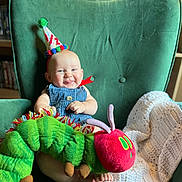 Christopher joined the competition — help win amazing prizes! baby, party_hat, plush_toy, caterpillar, green_armchair, knitted_blanket, smiling, tongue_out, indoor, child, cute, happy, seated, furniture, shelf, books, colorful, headwear, soft_toy, portrait