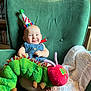 baby, party_hat, plush_toy, caterpillar, green_armchair, knitted_blanket, smiling, tongue_out, indoor, child, cute, happy, seated, furniture, shelf, books, colorful, headwear, soft_toy, portrait