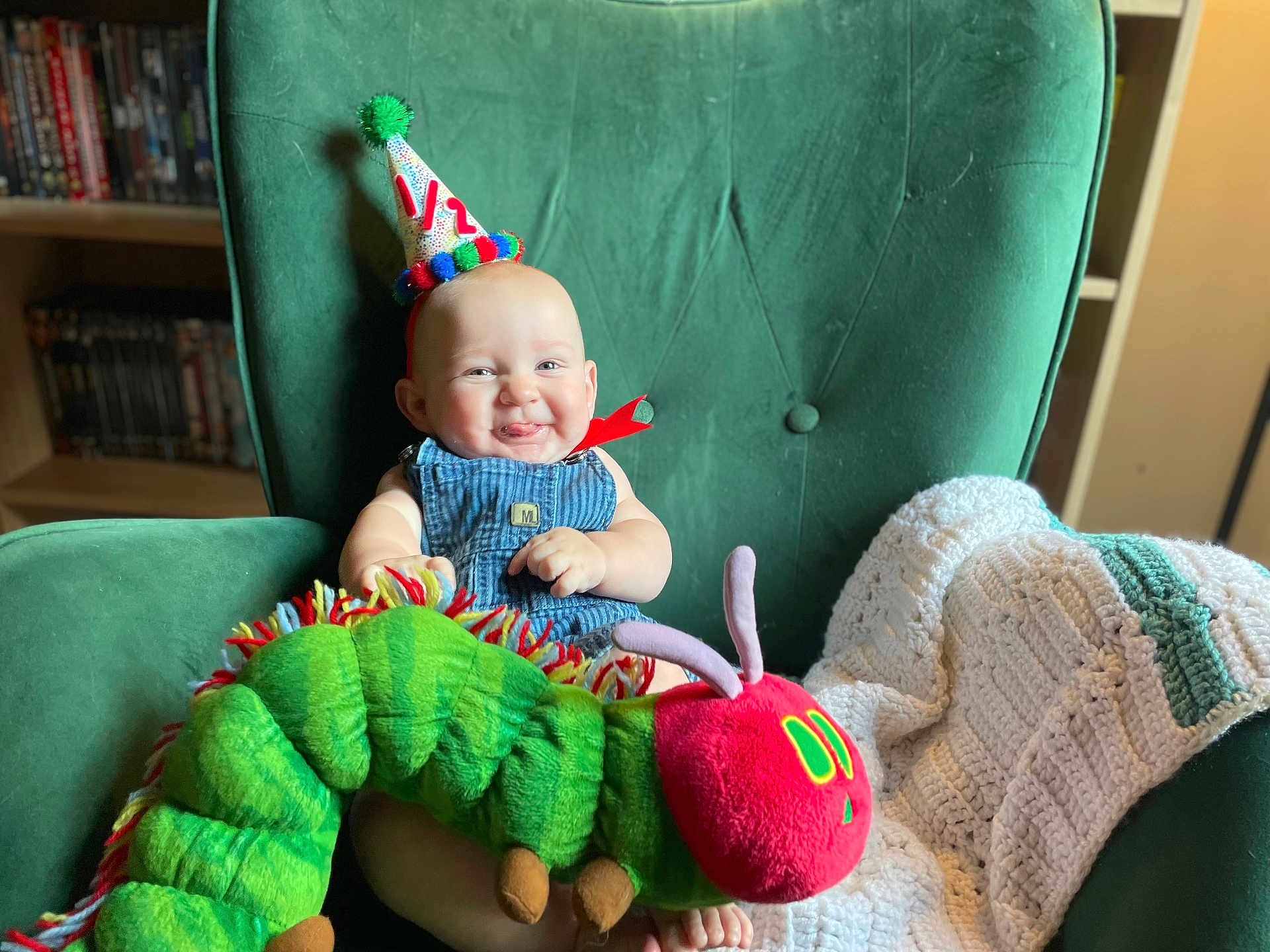 Christopher joined the competition — help win amazing prizes! baby, party_hat, plush_toy, caterpillar, green_armchair, knitted_blanket, smiling, tongue_out, indoor, child, cute, happy, seated, furniture, shelf, books, colorful, headwear, soft_toy, portrait