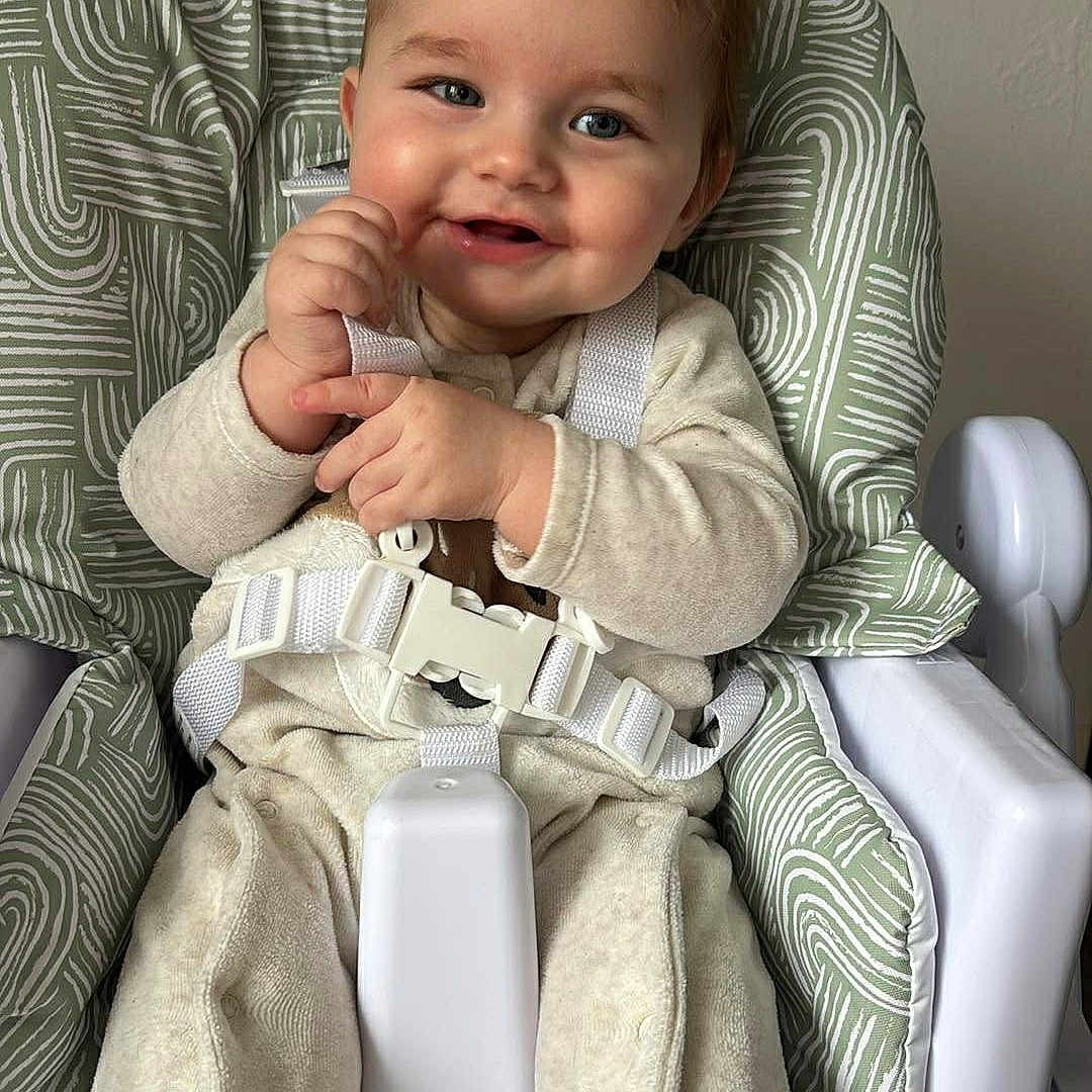 Ayden a rejoint le concours — aidez-le/la à gagner de superbes lots ! baby, child, high_chair, smile, face, infant, clothing, sitting, indoor, baby_outfit, safety_straps, happy, young_child, cute, person, furniture, patterned_fabric, neutral_background, light_skin, short_hair