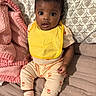 baby, child, yellow_bib, pink_blanket, patterned_pillow, bed, sitting, curious_eyes, cozy, indoors, clothing, sock, infant, cute, portrait, small_hands, looking_up, soft_texture, fabric, resting
