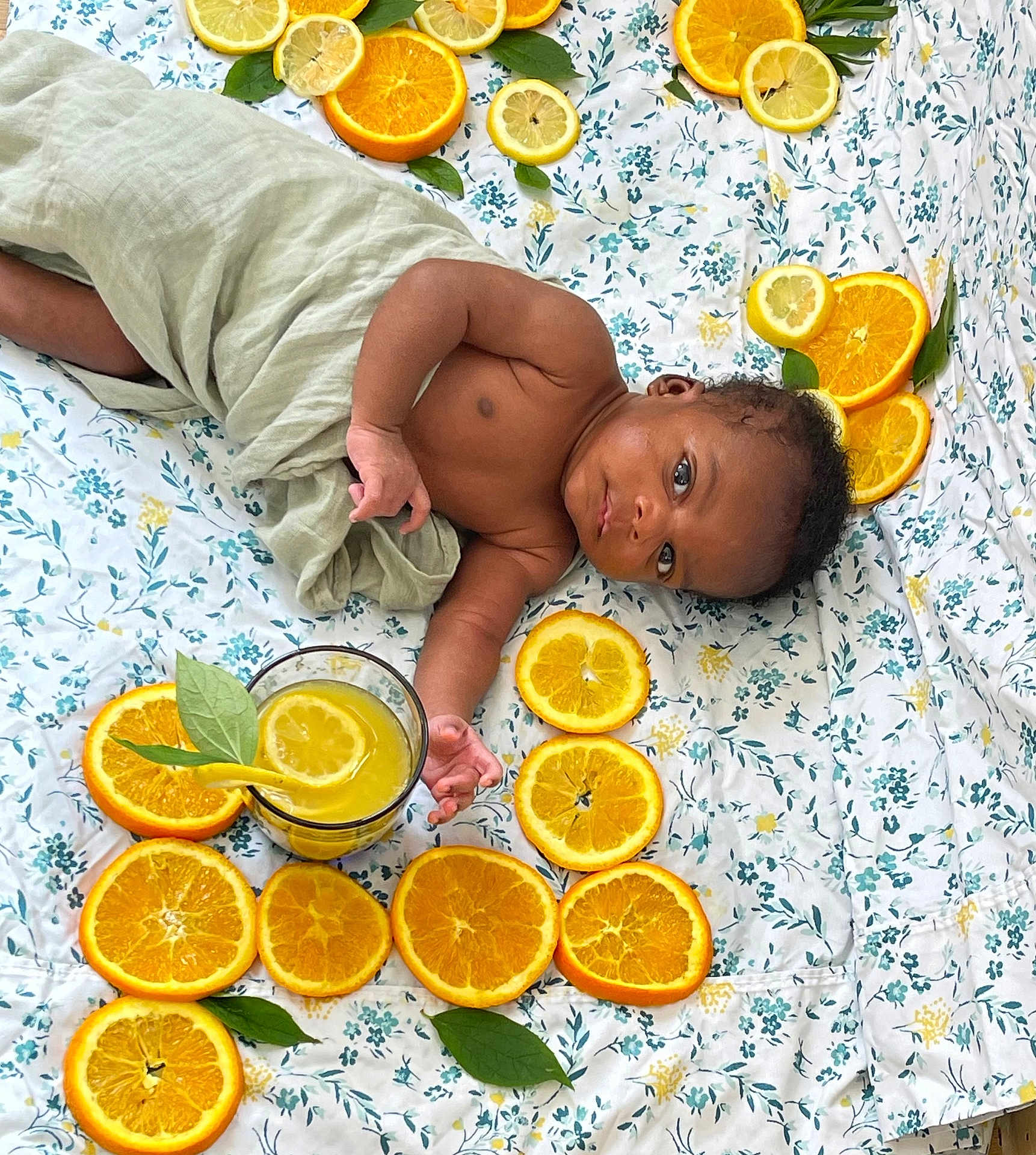 Zayden is registered to the contest to win money with this photo: baby, infant, orange_slices, lemon_slices, citrus, juice, glass, leaf, blanket, floral_pattern, wrapped, lying_down, curious, looking_at_camera, fresh, summer, fruit, drink, child, indoors