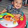 baby, toddler, child, smiling, indoor, walker, toy, steering_wheel, tiger_toy, batman_shirt, flooring, speaker, iron, table, plastic_toy, happy, person, face, clothing, playful