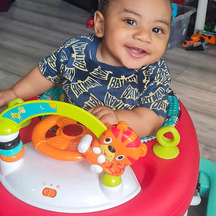 Kiemari is registered to the contest to win money with this photo: baby, batman_shirt, child, clothing, face, flooring, happy, indoor, iron, person, plastic_toy, playful, smiling, speaker, steering_wheel, table, tiger_toy, toddler, toy, walker