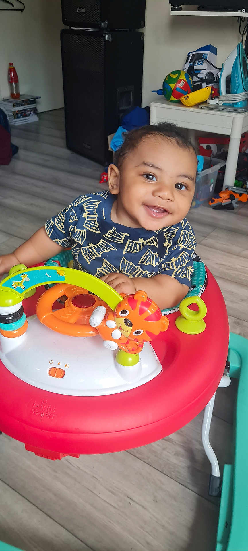 Kiemari is registered to the contest to win money with this photo: baby, toddler, child, smiling, indoor, walker, toy, steering_wheel, tiger_toy, batman_shirt, flooring, speaker, iron, table, plastic_toy, happy, person, face, clothing, playful
