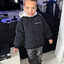 child, boy, indoor, jacket, hoodie, sneakers, jeans, living_room, television, game_controller, furniture, floor, standing, casual_clothing, smile, hair, person, modern, curious, coat