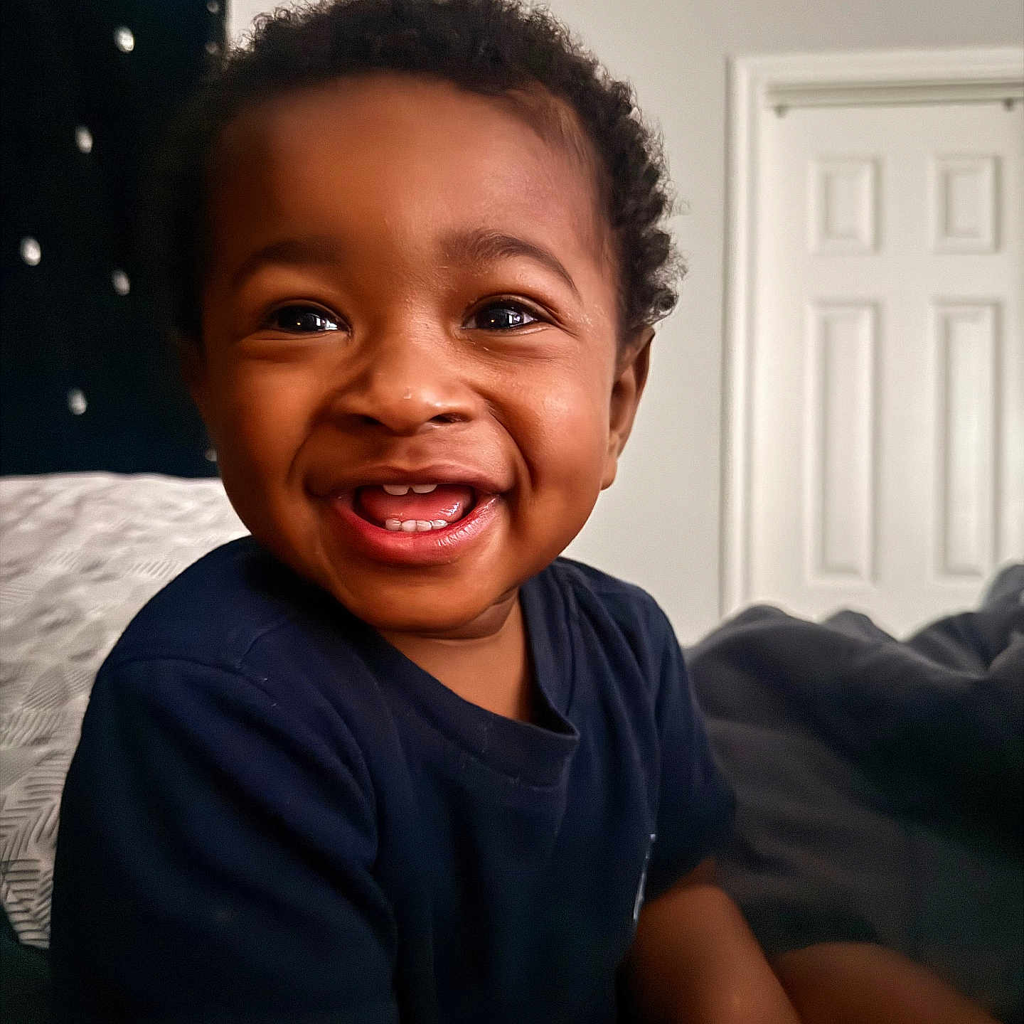 Willie is registered to the contest to win money with this photo: child, toddler, smiling, happy, indoor, bed, face, person, curly_hair, black_hair, dark_skin, clothing, blue_shirt, portrait, cute, young, sitting, home, door, cozy