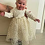 baby, child, dress, flower_headband, smiling, tongue_out, tattoo, hands, indoor, floor, wall, cute, person, portrait, white, lace, happy, infant, young, standing