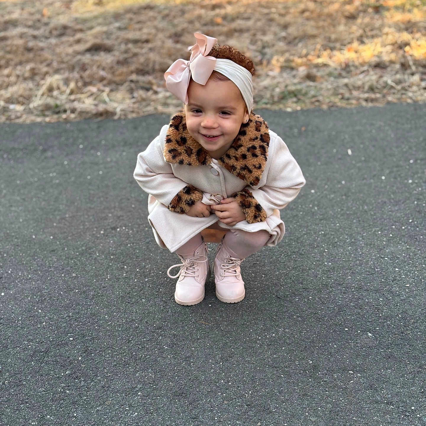 Avianna is registered to the contest to win money with this photo: boots, bow, child, coat, crouching, curly_hair, cute, daylight, fashion, grass, headband, leopard_print, nature, outdoor, pavement, person, playful, smile, toddler, young_child