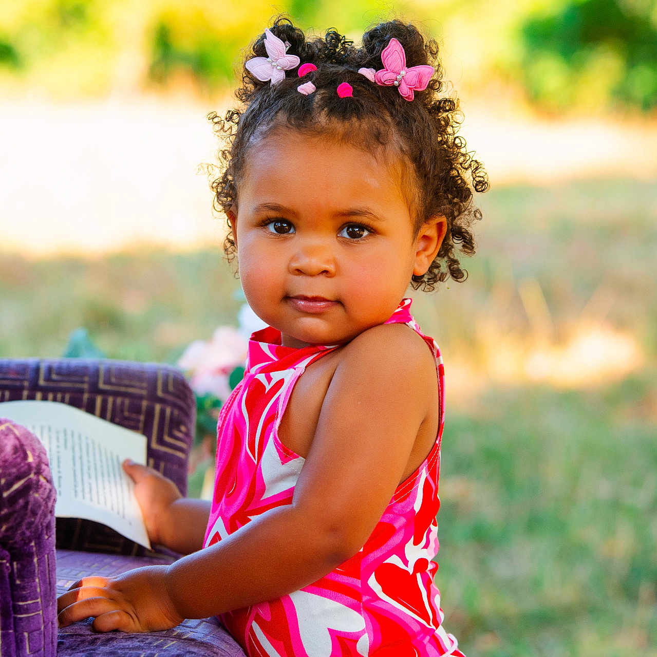 Layana a rejoint le concours — aidez-le/la à gagner de superbes lots ! book, butterfly_clips, child, curly_hair, cute, daylight, grass, hair_clips, nature, outdoor, person, pink_dress, playful, portrait, purple_chair, reading, smiling, summer, toddler, young_child