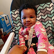 Lilliana is registered to the contest to win money with this photo: baby, child, high_chair, pink_onesie, curly_hair, holding_hand, indoor, patterned_chair, smiling, person, infant, cute, hand, furniture, room, casual, comfort, baby_clothes, toddler, care