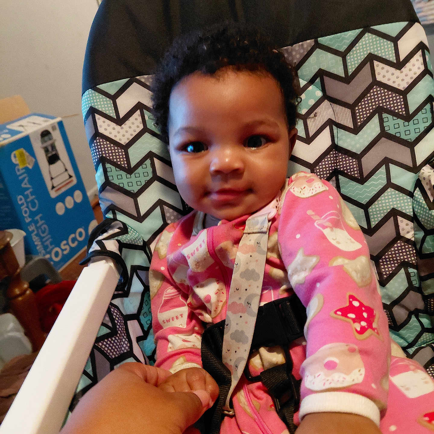 Lilliana is registered to the contest to win money with this photo: baby, baby_clothes, care, casual, child, comfort, curly_hair, cute, furniture, hand, high_chair, holding_hand, indoor, infant, patterned_chair, person, pink_onesie, room, smiling, toddler