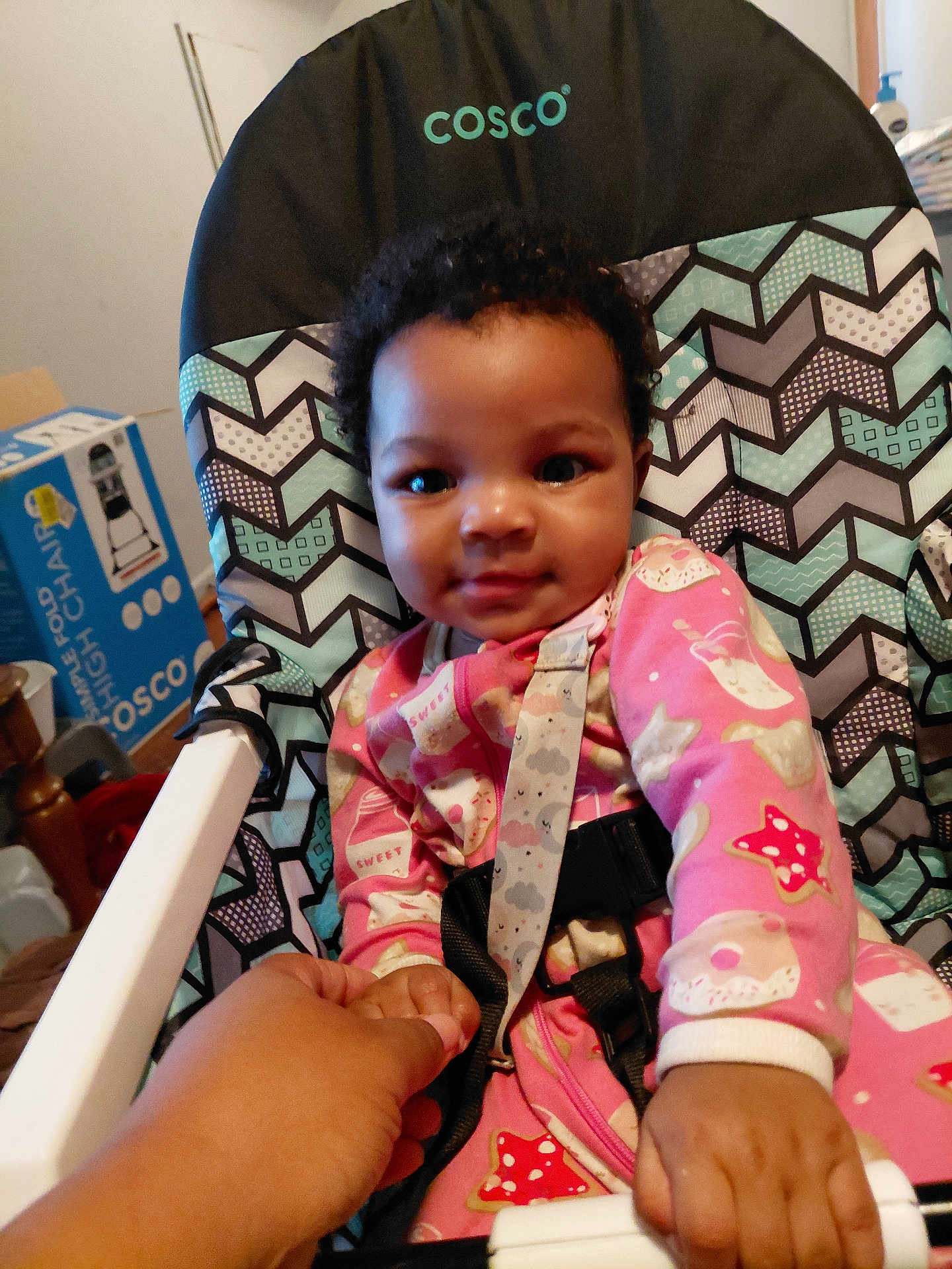 Lilliana is registered to the contest to win money with this photo: baby, child, high_chair, pink_onesie, curly_hair, holding_hand, indoor, patterned_chair, smiling, person, infant, cute, hand, furniture, room, casual, comfort, baby_clothes, toddler, care