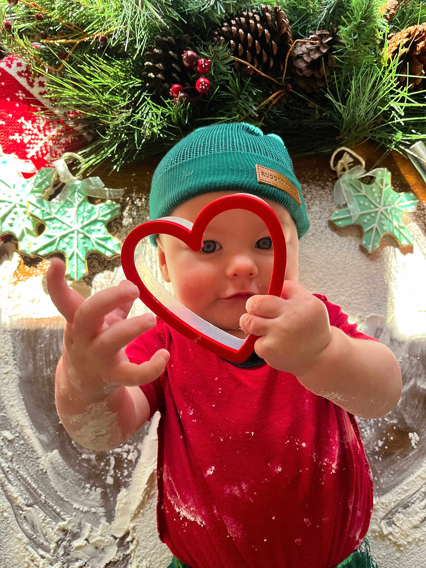 Ronan joined the competition — help win amazing prizes! toddler, child, flour, cookie_cutter, heart_shape, red_shirt, green_beanie, holiday_decor, pinecones, greenery, baking, kitchen, playful, hands, face, curious, festive, christmas, cookies, indoors