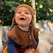 Ronan is registered to the contest to win money with this photo: baby, child, smiling, hat, knit_hat, brown_clothing, bib, hands, floor, indoor, festive_lights, blurred_background, greenery, happy, cute, portrait, infant, warm_clothing, candid, closeup