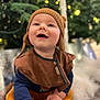 baby, child, smiling, hat, knit_hat, brown_clothing, bib, hands, floor, indoor, festive_lights, blurred_background, greenery, happy, cute, portrait, infant, warm_clothing, candid, closeup
