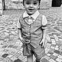 child, boy, cap, bow_tie, vest, shorts, sandals, cobblestone, street, stone_building, smiling, portrait, standing, outdoor, monochrome, black_and_white, cute, fashion, happy, person