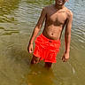 boy, child, water, swimming_trunks, outdoor, nature, pond, lake, summer, sunlight, trees, sandy_bank, greenery, standing, casual, daytime, water_body, young, barefoot, squinting