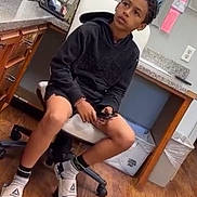Kaide is registered to the contest to win money with this photo: child, boy, indoor, sitting, chair, black_hoodie, shorts, socks, slides, phone, office, desk, trash_bin, wood_floor, wall_outlet, paper, container, curly_hair, looking_away, casual_clothing