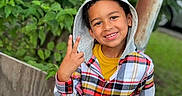 Kaide is registered to the contest to win money with this photo: child, smiling, hoodie, plaid_shirt, outdoor, greenery, peace_sign, casual_clothing, happy, portrait, nature, person, grass, wooden_fence, young, cute, playful, tree, daylight, casual