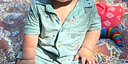 Jameson is registered to the contest to win money with this photo: baby, child, outdoor, blanket, patterned_blanket, clothing, shorts, shirt, face, expression, sunlight, grass, picnic, toy, colorful, sitting, legs, feet, hand, skin