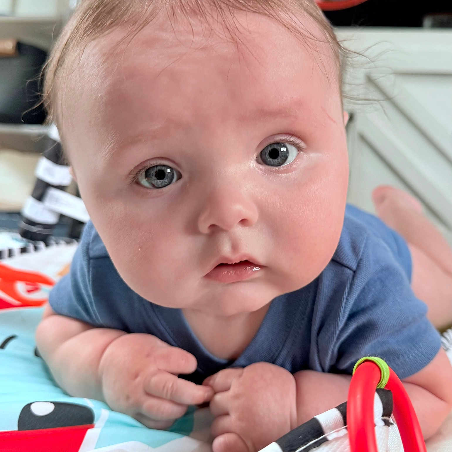 Jameson is registered to the contest to win money with this photo: baby, blue_shirt, child, closeup, colorful, curious, cute, expression, eyes, face, hands, indoors, infant, laying, playmat, portrait, red_ring, skin, toy, young