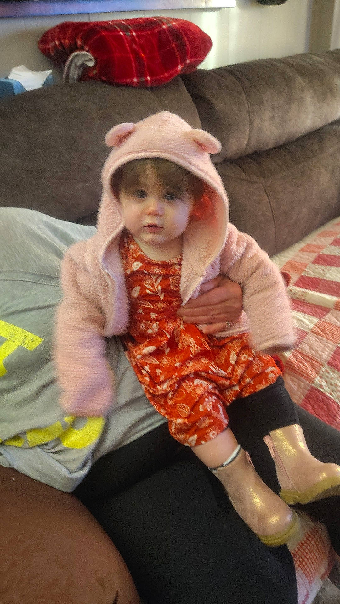Paisley is registered to the contest to win money with this photo: baby, blanket, boots, brown_furniture, casual_clothing, child, couch, cozy, cute, hand, home, hood, indoor, jacket, orange_dress, person, plaid, portrait, sitting, toddler