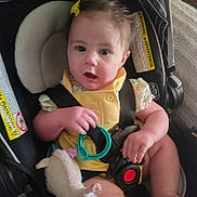 Paisley is registered to the contest to win money with this photo: baby, infant, car_seat, yellow_clothing, toy, plush_toy, child, sitting, straps, safety_harness, indoor, curly_hair, expression, hand, foot, warning_label, seat, comfort, cute, child_care