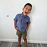 child, boy, smiling, laughing, portrait, standing, indoors, shirt, shorts, sandals, wall, floor, wood_floor, home, playful, happy, face, eyes, hair, full_body