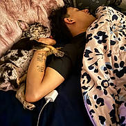 Rallo is registered to the contest to win money with this photo: person, dog, sleeping, bed, blanket, paw, tattoo, fur, pillow, resting, cozy, indoor, black_shirt, patterned_blanket, charging_cable, relaxed, companion, spot_pattern, cute, nap