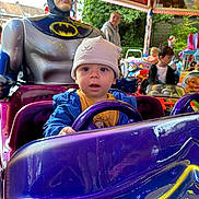 Isaac participe au concours pour gagner de l'argent avec cette photo : toddler, child, batman, ride, amusement_park, toy_car, hat, blue_jacket, steering_wheel, statue, adult, smile, outdoor, colorful, lights, fun, playful, background, people, park